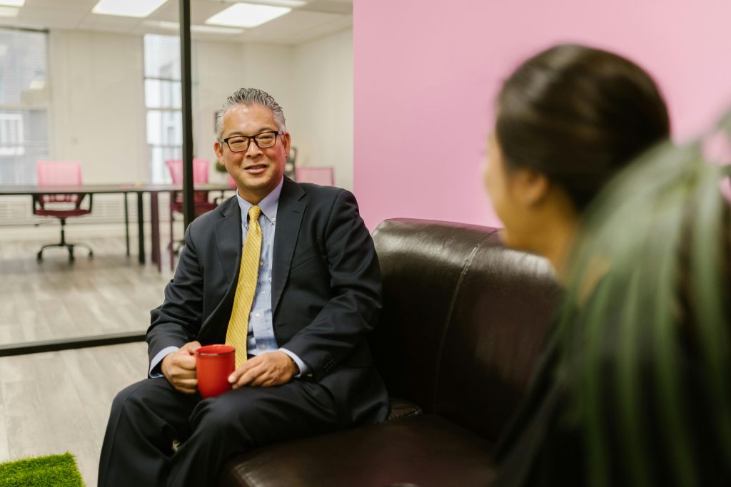 A businessman in black suit Sitting on leather couch in an office, drinking a coffee, smiling and talking to a co-worker